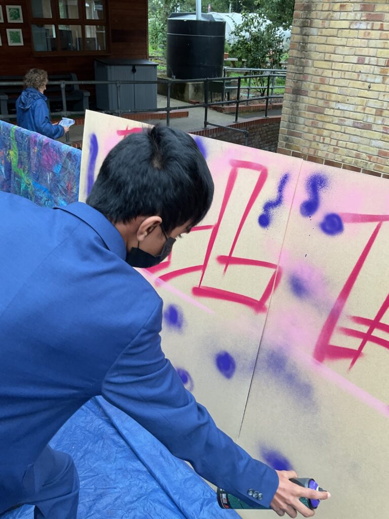 A person in a blue suit jacket and a face mask is bending down and using a can of magenta spray paint to create graffiti art on a large, light-colored board outdoors. Some magenta and purple markings are already visible on the board. Another person is visible in the background.
