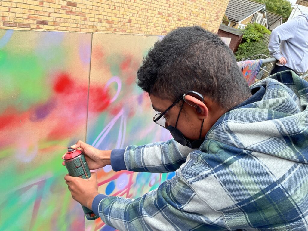 A person wearing a plaid hooded shirt and a face mask is closely applying red spray paint to a section of the art board. The board already has various colors like green, red, and purple on it.