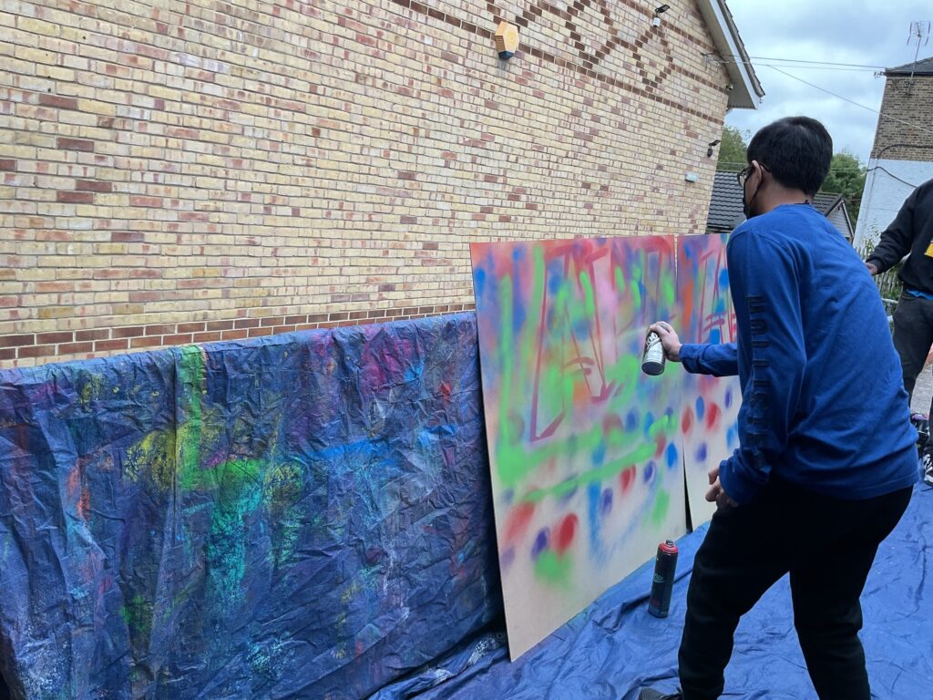 A person in a long-sleeved blue shirt and a face mask is standing and using a can of white spray paint on the outdoor art board, which is already covered in a vibrant mix of colors and designs. A large blue tarp is on the ground.
