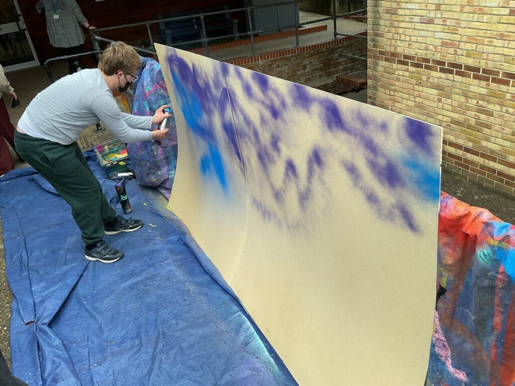 A person in a grey long-sleeved shirt, dark trousers, and a face mask is using a can of spray paint to apply blue and purple colors to a large, light-colored board outdoors. The board is positioned against a brick wall, and a blue tarp is on the ground.
