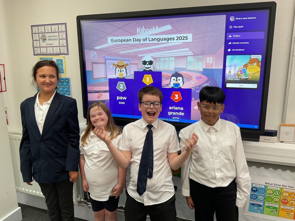 This image shows four children celebrating their success in a Kahoot! quiz on the topic of "European Day of Languages 2025". They are standing in front of a large screen displaying the quiz results, with usernames like "paw" and "ariana grande" and scores. The children appear excited; the boy in the center with glasses is particularly expressive, throwing his hands up. They are all wearing uniform-style clothing (shirts, ties, and blazers), suggesting a formal school environment.