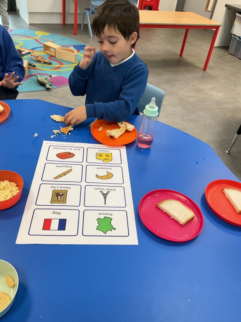 This image shows a young boy participating in a cultural or language-themed snack time activity, specifically related to France. The boy is sitting at a blue table, eating bread. In front of him is a worksheet with pictures and words related to French culture and food, including: Flag, France (map), Mona Lisa, Eiffel Tower, Pain au chocolat, Cheese, Baguette, and Croissant. He has slices of bread and a small amount of crumbles on his plate, likely being used to learn about or taste the different items listed on the sheet.