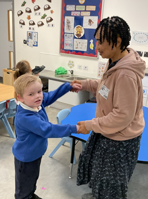 An adult woman and a young boy wearing a blue school jumper are standing in a classroom. They are holding hands, facing each other, and appear to be dancing or playing a game. The classroom has decorations on the wall and storage cabinets.