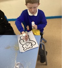 A young boy wearing a blue school jumper is seated at a table, focusing on applying glue or glitter to a printed outline drawing of Pudsey Bear. There is spilled gold glitter on the blue table surface and he holds a bottle of glitter in each hand.