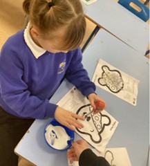 A young girl wearing a purple school jumper is seated at a desk, carefully applying glue from a bottle onto a printed outline of Pudsey Bear. A small bowl of white adhesive is also on the desk.