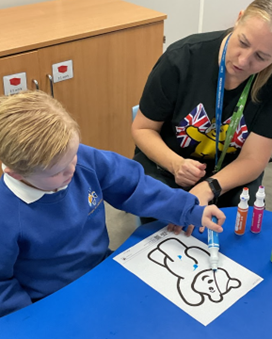 A young boy in a blue school jumper is seated at a blue table, working on a coloring activity of Pudsey Bear. An adult woman with blonde hair, wearing a black shirt with a British flag design, is leaning over to guide him as he uses a colorful marker pen on the paper.