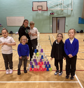 A group of five smiling young children and two adults pose in a gym next to a very tall, multi-colored pyramid made of stacked plastic cups.