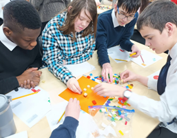 An overhead view of a group of four students (two boys and two girls) gathered around a table, actively engaged in building with colorful plastic bricks (similar to LEGO). They are focused on the small structures they are creating on an orange baseplate, surrounded by loose bricks, paper, and pencils. One student's hand is visible in the foreground.