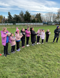 : A line of about ten young girls and one adult standing on a grass field, all holding rugby balls, seemingly listening to instruction from a second adult on the far right.