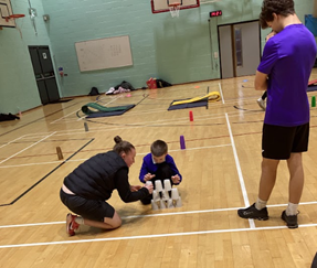 An adult kneels next to a child who is concentrating on carefully placing a clear plastic cup on top of a small cup pyramid they are building on a wooden gym floor. An older student watches from the right.