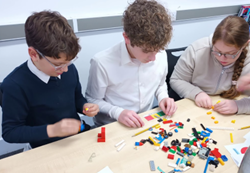A medium shot of three students (two boys and one girl) sitting side-by-side at a wooden table, also building with colorful plastic bricks. They are concentrated on manipulating the bricks scattered on the table in front of them, each working on their own or a shared small construction.