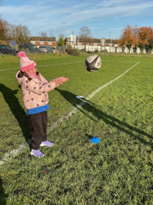 A young girl, wearing a pink bobble hat and jacket, standing on a grass field and reaching out to catch a rugby ball that is suspended in the air.