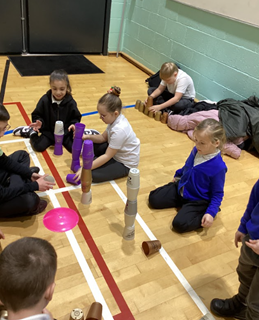 Several children are seated on a gym floor, engaged in different building activities, including stacking purple plastic cups into tall towers and arranging small terracotta pots.