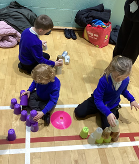 Several children are seated on a gym floor, engaged in different building activities, including stacking purple plastic cups into tall towers and arranging small terracotta pots.