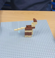 A close-up image of a small structure built with brown and tan plastic bricks on a grey baseplate. The structure appears to represent an animal or object with a body, head, and an elongated tail-like piece.
