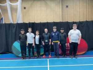 A group portrait of six children and one adult standing in a line inside an indoor sports hall. They are posed behind two curved "Active" branded banners on a blue court with a high brick wall in the background.