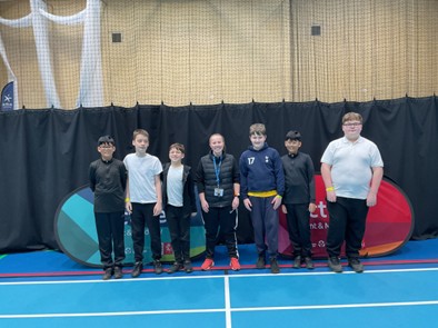 A group portrait of six children and one adult standing in a line inside an indoor sports hall. They are posed behind two curved "Active" branded banners on a blue court with a high brick wall in the background.