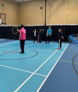 An indoor sports hall scene where a person wearing a pink "Events Team" t-shirt holds up a yellow hoop. A group of children and a coach stand nearby on the blue court, which is marked with various sport lines.