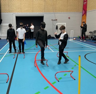 Two children wearing blindfolds navigate an obstacle course in a gymnasium. They are stepping over small red hurdles on a blue floor while other children and sports equipment, like cricket stumps and cones, are visible in the background.