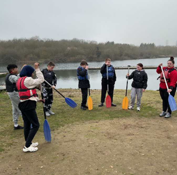 A group of young people and an instructor stand in a semi-circle on a grassy bank by a lake, holding colorful paddles and wearing life jackets.