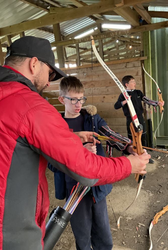 An instructor in a red jacket and black cap assists a boy in a navy jacket with an archery bow in an indoor range.