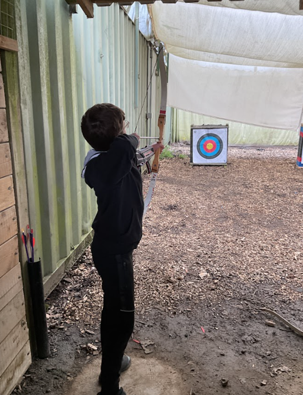 A young person in a black hoodie stands in an archery range, aiming a bow and arrow at a colorful target in the distance.