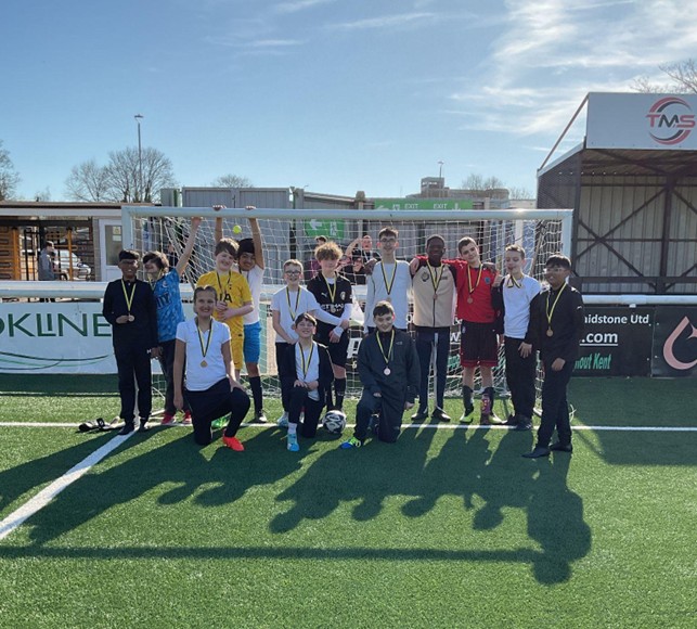 A different group of seven young people standing in a line inside a football goal. Most are wearing medals, and a football sits on the grass at their feet.