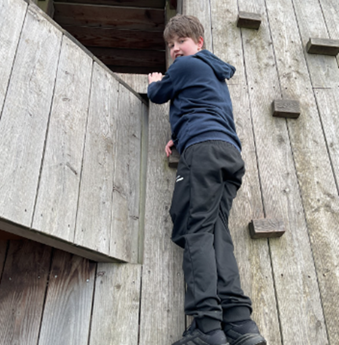 A boy in a navy hoodie and black trousers climbs a wooden climbing wall, looking back over his shoulder toward the camera.