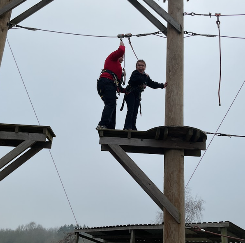 An instructor and a young person, both in harnesses, navigate a high-ropes course on a wooden platform high above the ground.