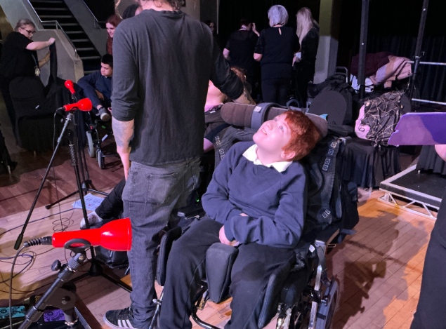 A smiling person in a wheelchair looks upwards while on a stage, with someone standing beside them and red-covered microphone stands positioned nearby.