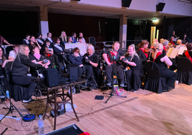 Several individuals in wheelchairs are positioned at the front of a stage, supported by staff members and using small electronic devices, with an audience and an orchestra seated behind them.