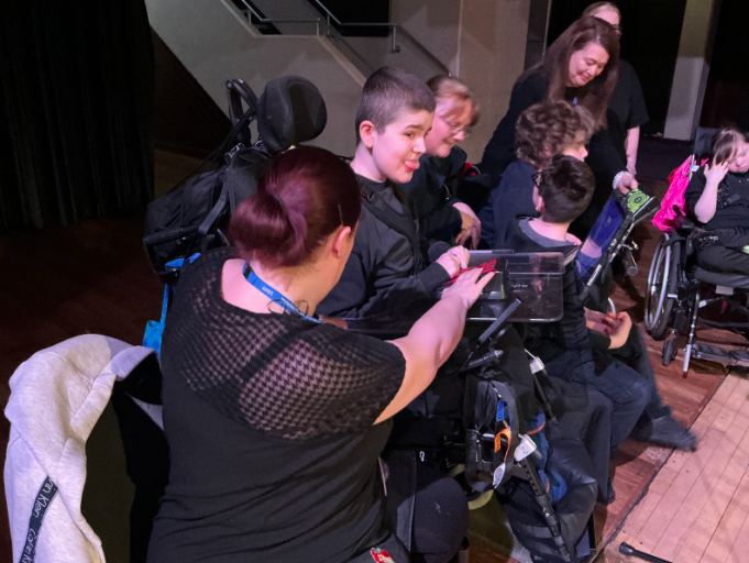 A staff member leans in to assist a person in a wheelchair who is pressing a button on a flat electronic device resting on their tray, with other attendees gathered behind them.