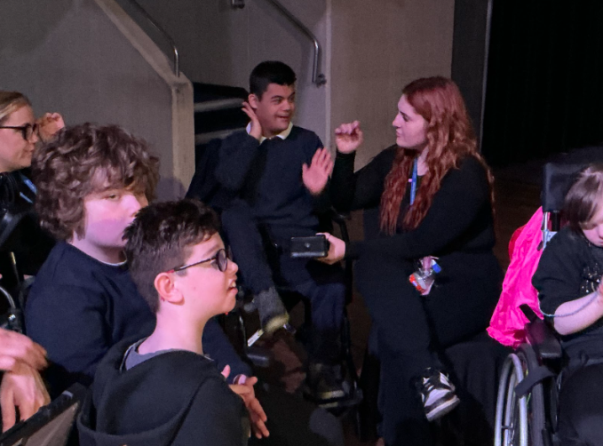 A staff member interacts with a seated person, using hand gestures, while others, including individuals in wheelchairs, sit nearby in an auditorium setting.