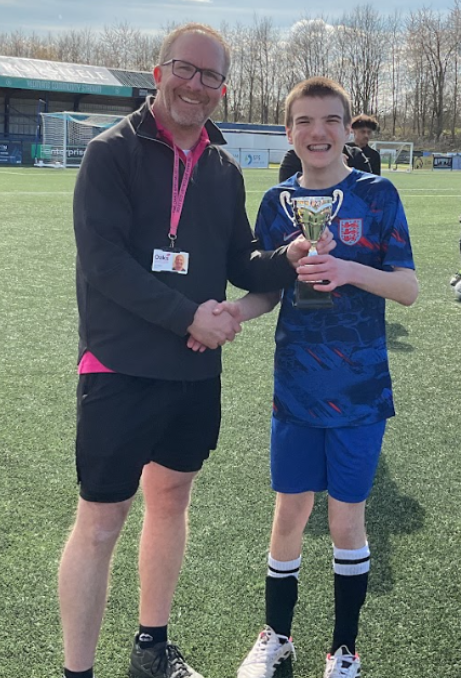 A young football player in a blue kit receives a trophy from an adult.