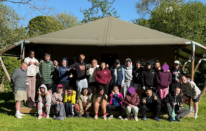 A group of students and teachers pose for a photo in an outdoor setting in front of a large green tent-like building