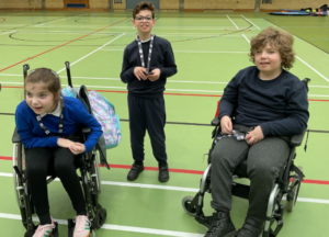 Three children smiling in an indoor sports hall. Two of the children are seated in wheelchairs, and one is standing in the middle.