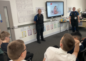 Two police officers standing in a classroom discussing online safety with students. The TV screen is displaying a picture of a girl on a phone