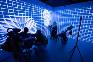 A wide shot of an immersive sensory room with walls covered in a glowing blue grid projection, as children in wheelchairs and adults interact with the light displays.