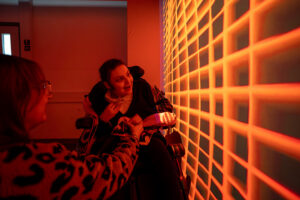 A young girl in a wheelchair smiles at a woman, cast in a vibrant red and orange glow from a geometric grid pattern projected onto the wall.