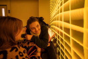 A smiling young girl in a wheelchair looks at a woman in a leopard-print jacket, both illuminated by a warm yellow grid pattern projected onto a large wall.