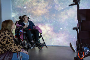 A young girl in a wheelchair and a caregiver look up at a large, immersive wall projection of a colorful cosmic nebula.
