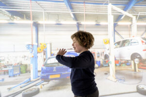 A young boy looking excited with his hands raised stands in front of a large wall projection depicting the inside of a car repair garage.