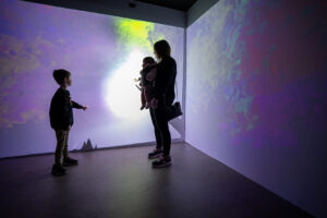 Silhouette of a woman holding a baby in a carrier alongside a young boy pointing at an immersive, colorful wall projection of swirling purple, green, and white light.